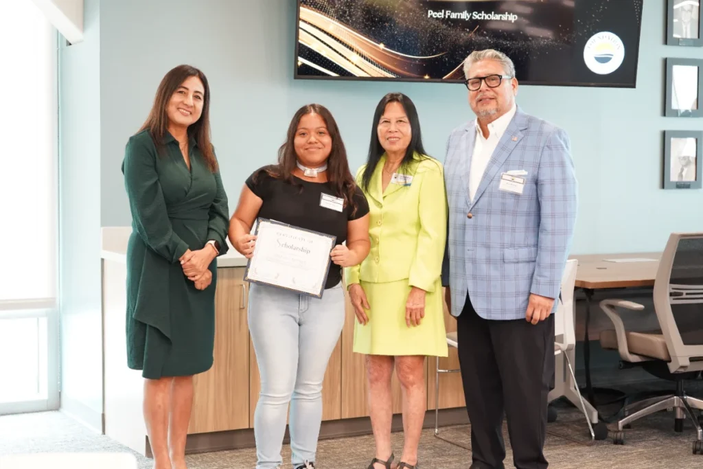 Four people stand indoors; the second from the left holds a scholarship certificate. A screen in the background displays "Pool Family Scholarship.