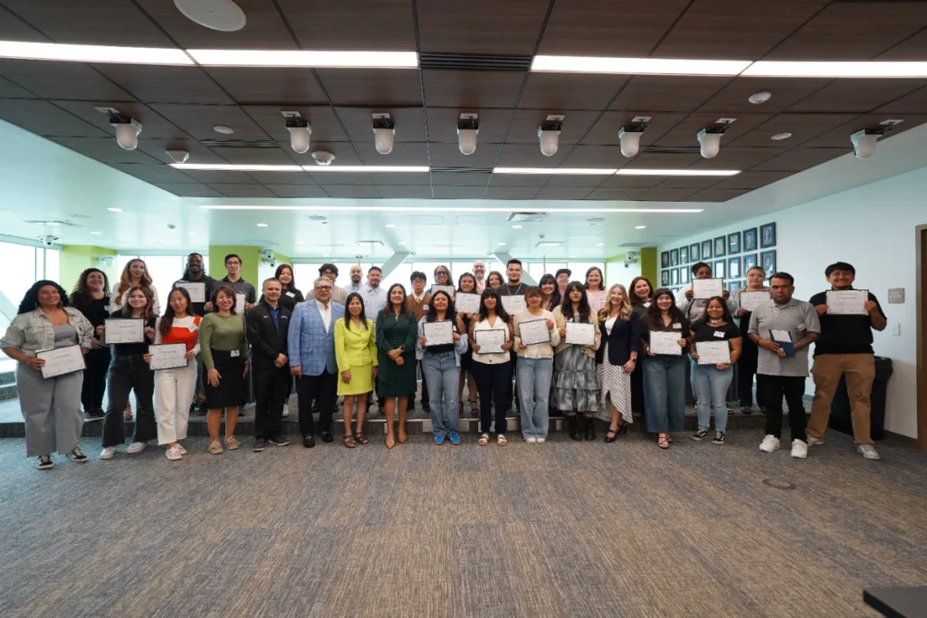 A large group of people stands indoors, some holding certificates, posing for a group photo in a well-lit room with plaques on the wall.