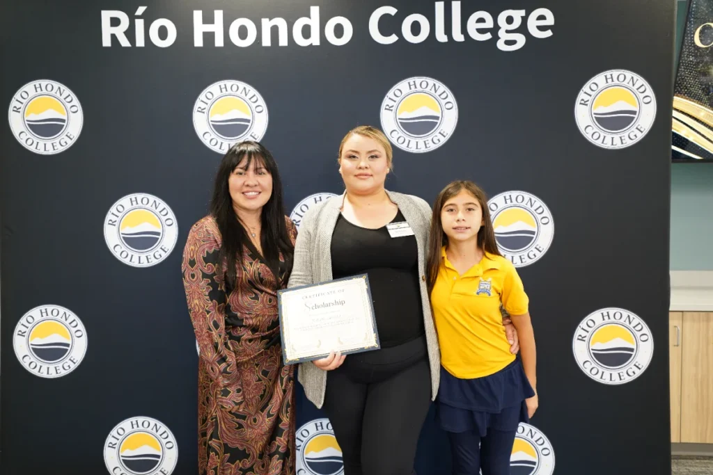 Three people stand in front of a Río Hondo College backdrop; the person in the center holds a certificate.