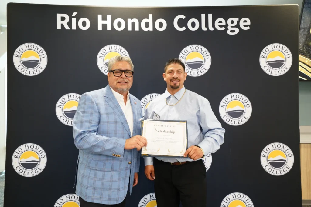 Two men stand in front of a “Río Hondo College” backdrop. One is holding a scholarship certificate and both are smiling at the camera.