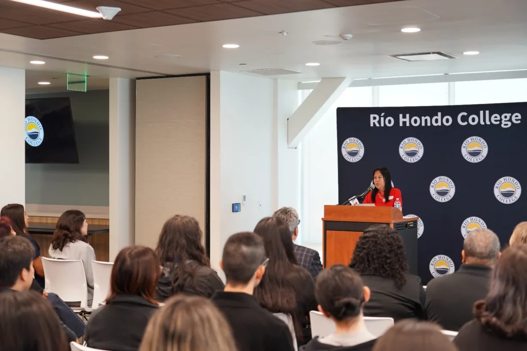 A woman speaks at a podium in front of an audience at Río Hondo College, with a branded backdrop behind her.