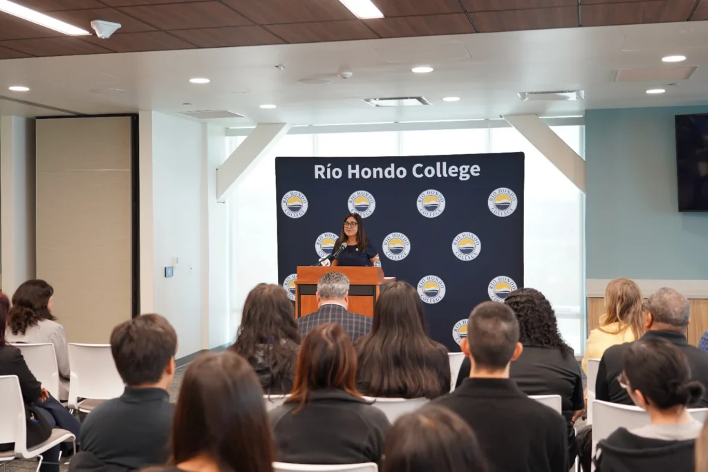 A woman stands at a podium speaking to an audience in a room with a "Río Hondo College" banner in the background.