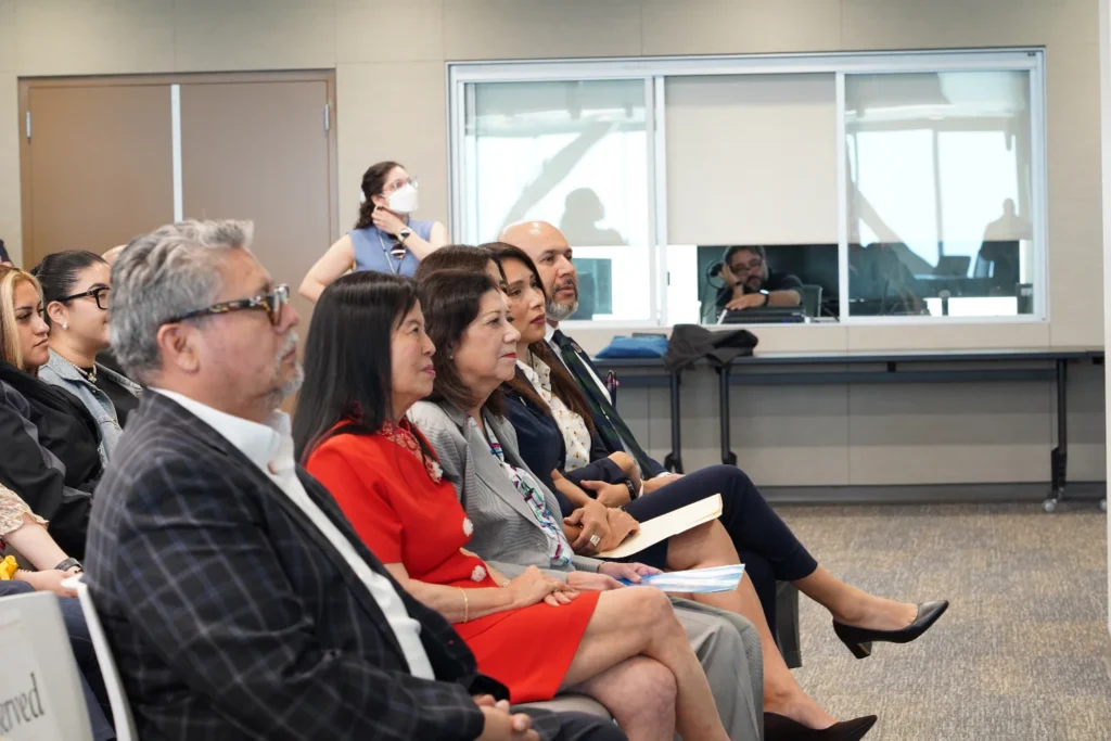 A group of people sits in chairs, attentively listening to a presentation in a conference room; one person in the background wears a mask.
