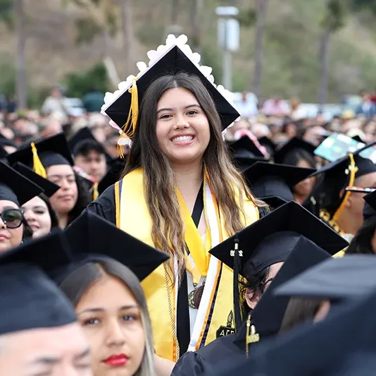 A smiling graduate in cap and gown stands among a crowd of fellow graduates at the 63rd Commencement outdoor ceremony.