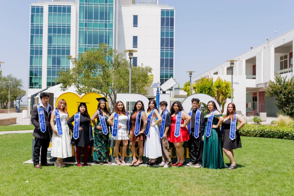 A group of graduates wearing sashes and formal attire stand in front of a large campus sign on a grassy area with modern buildings in the background.
