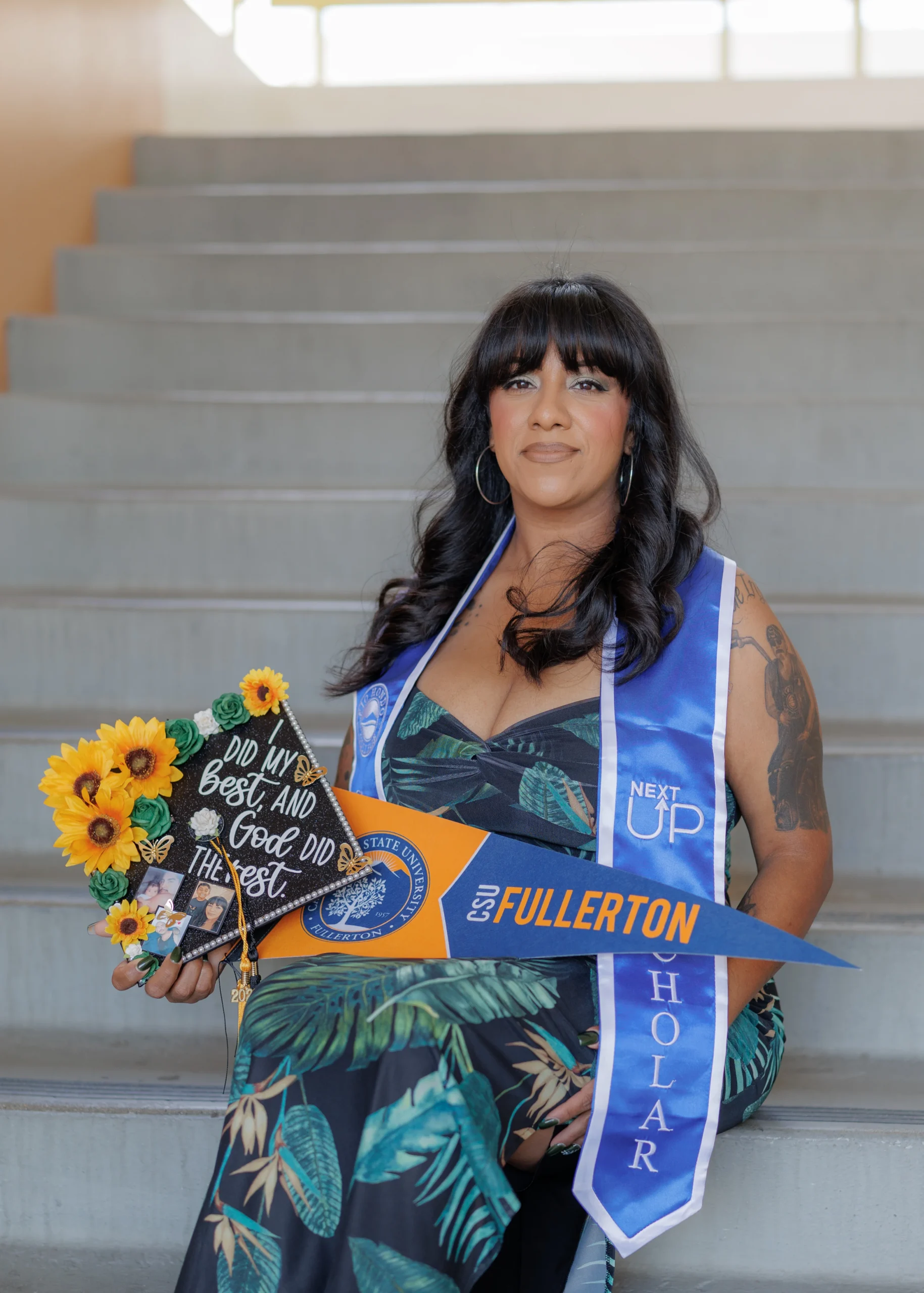 A woman in a floral dress sits on stairs holding a decorated graduation cap and a Cal State Fullerton pennant, wearing a Next Up honor stole and a graduation stole.