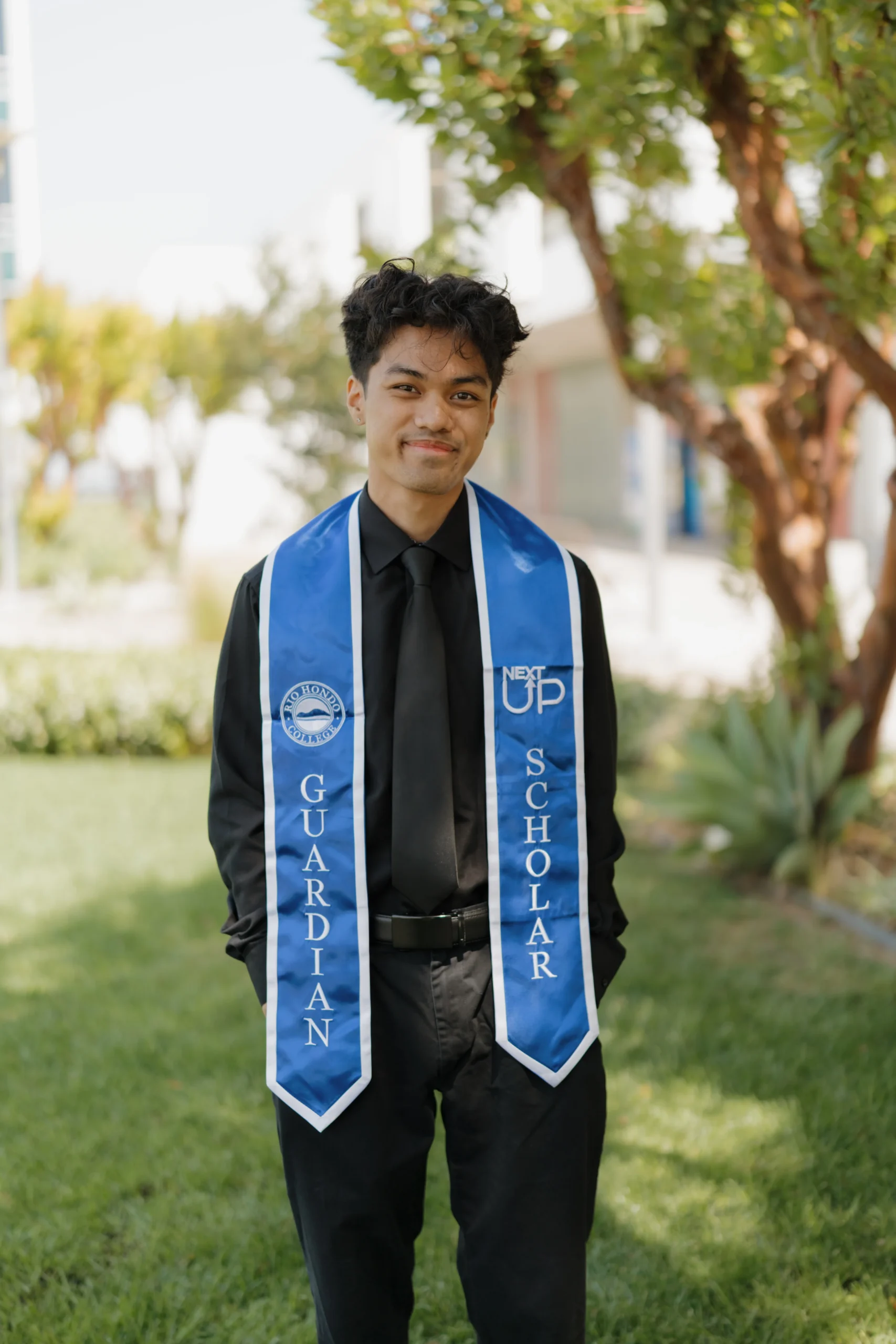 A young person in a black shirt and tie stands outside, wearing a blue graduation stole that reads "Guardian Scholar" and "Next Up.