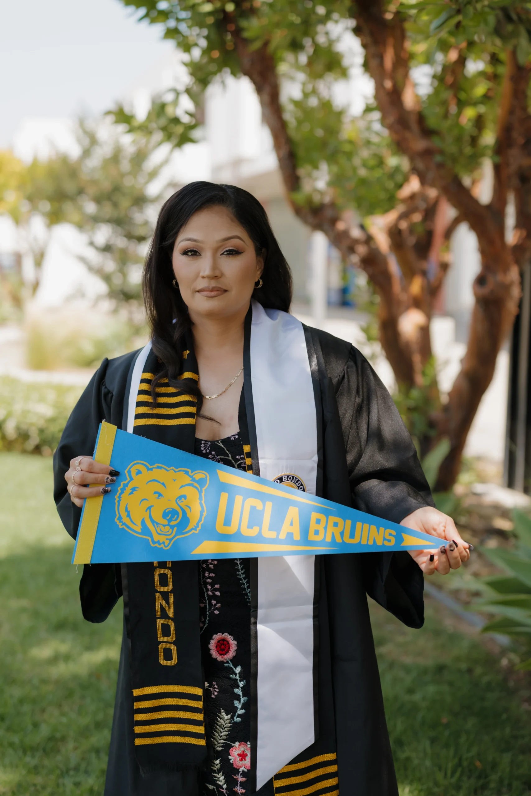 A person in a graduation gown holds a UCLA Bruins pennant while standing outside on a sunny day.
