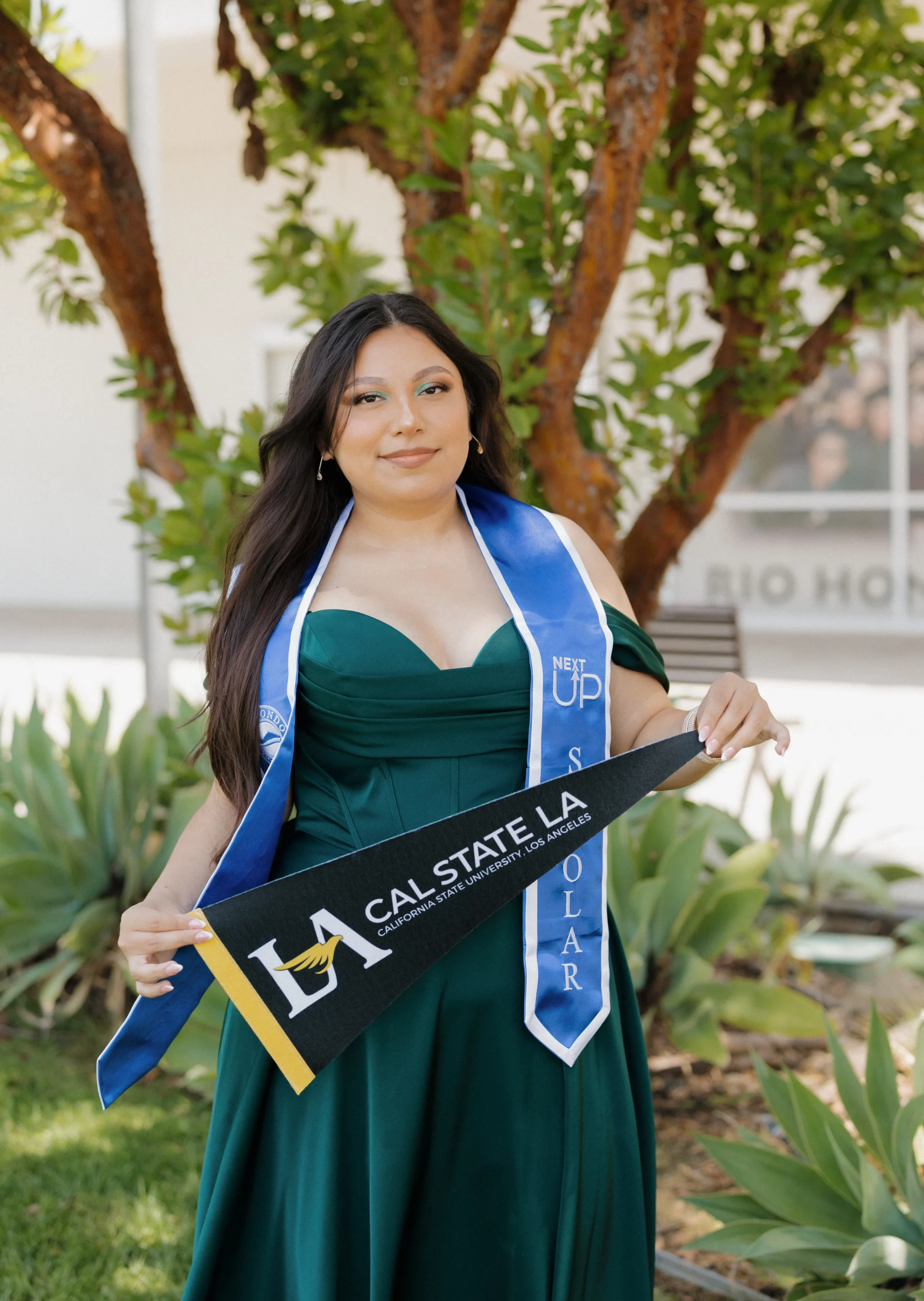 A woman in a green dress stands outdoors, wearing graduation stoles and holding a Cal State LA pennant.