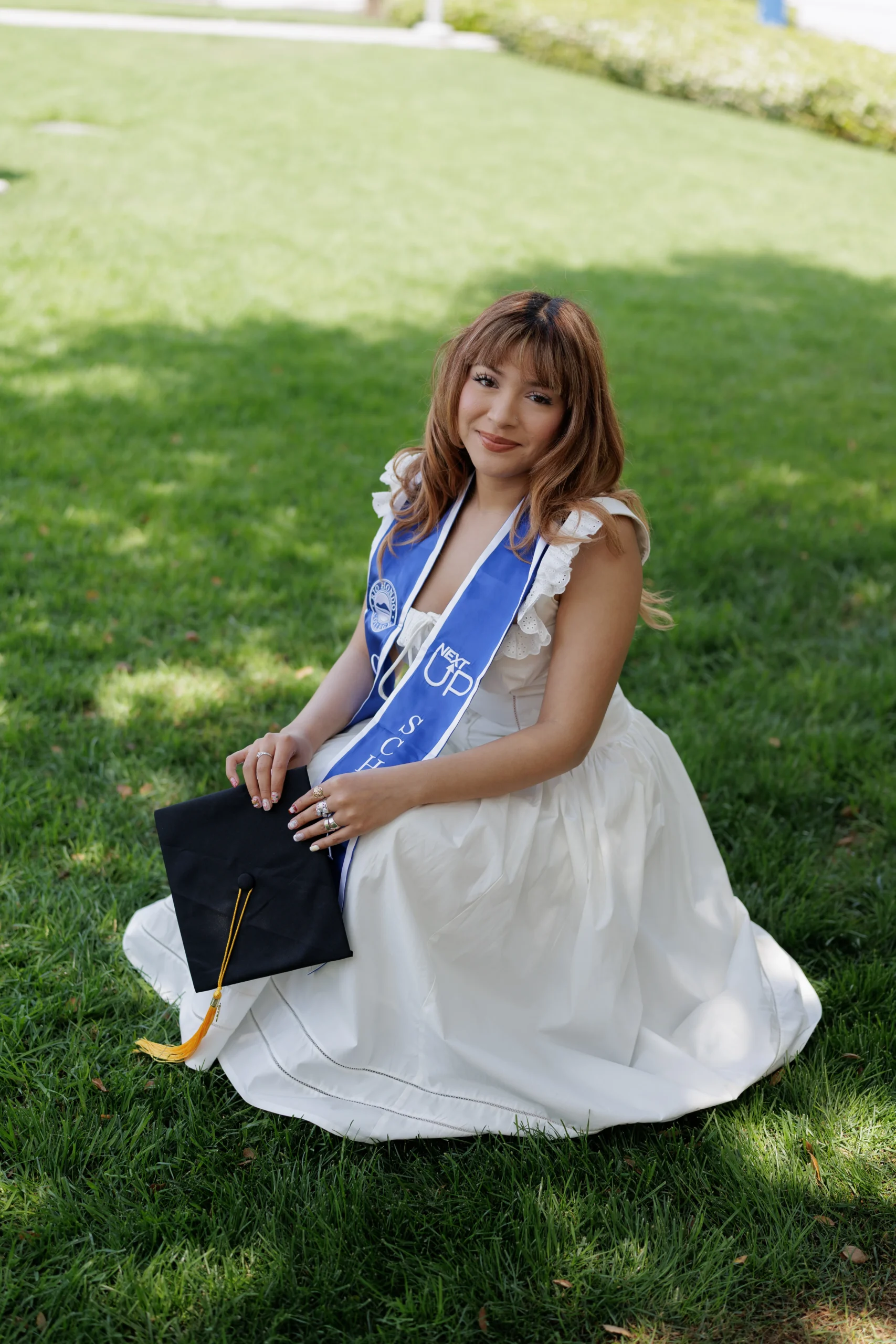 A young woman in a white dress and blue graduation stole sits on grass holding a graduation cap.