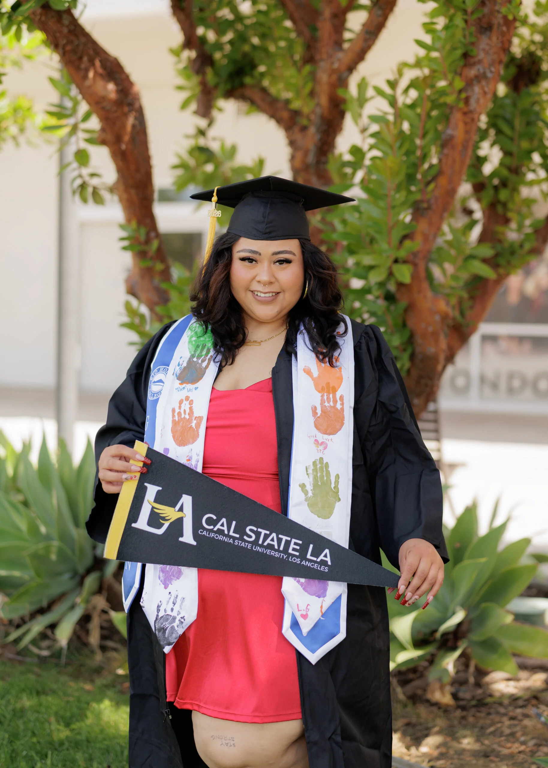 A graduate in a cap and gown holds a "Cal State LA" pennant and smiles outdoors, wearing a red dress and a stole with colorful handprints.