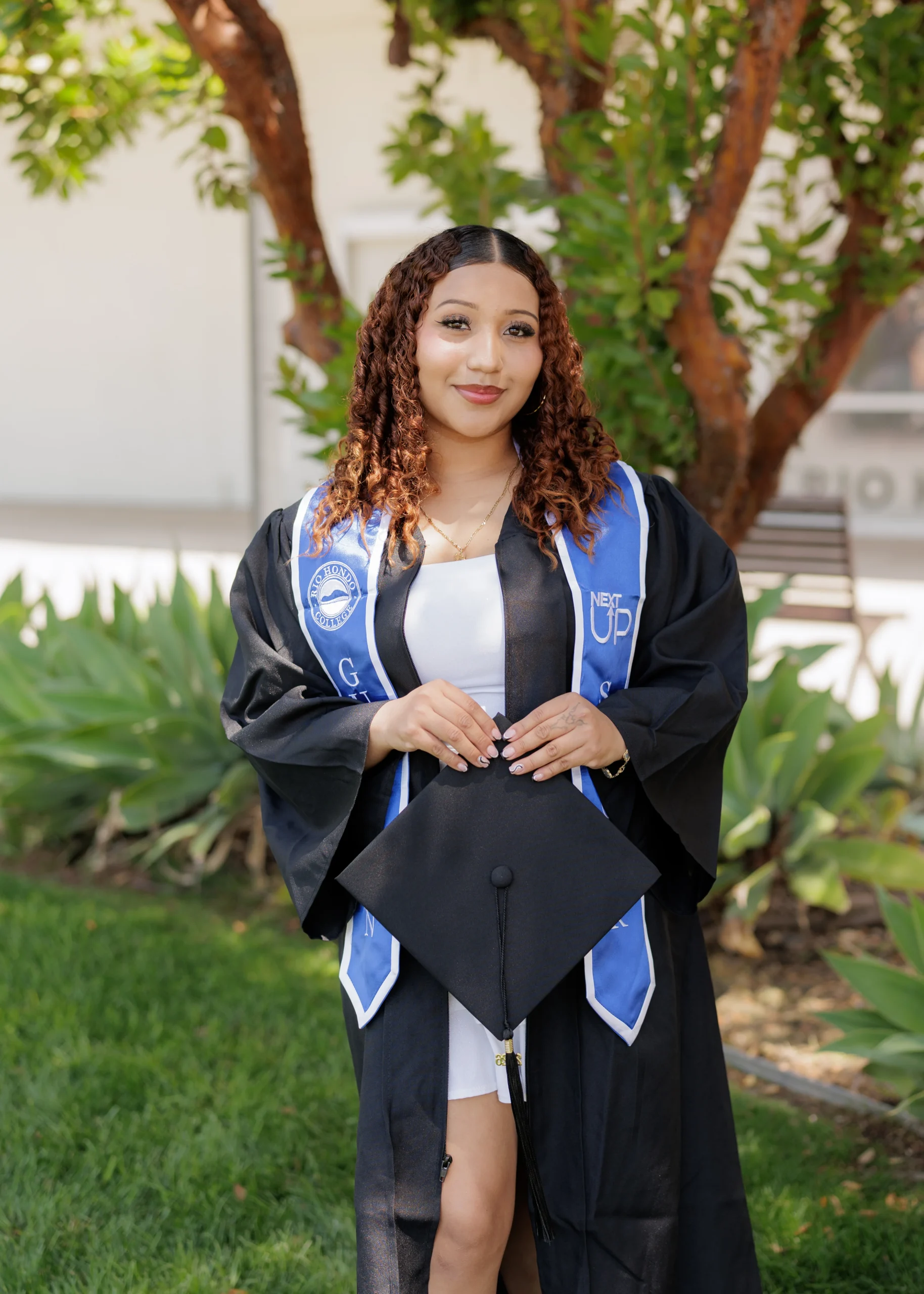 A young woman in a graduation gown and stole holds her cap, standing outside in front of greenery and a tree.
