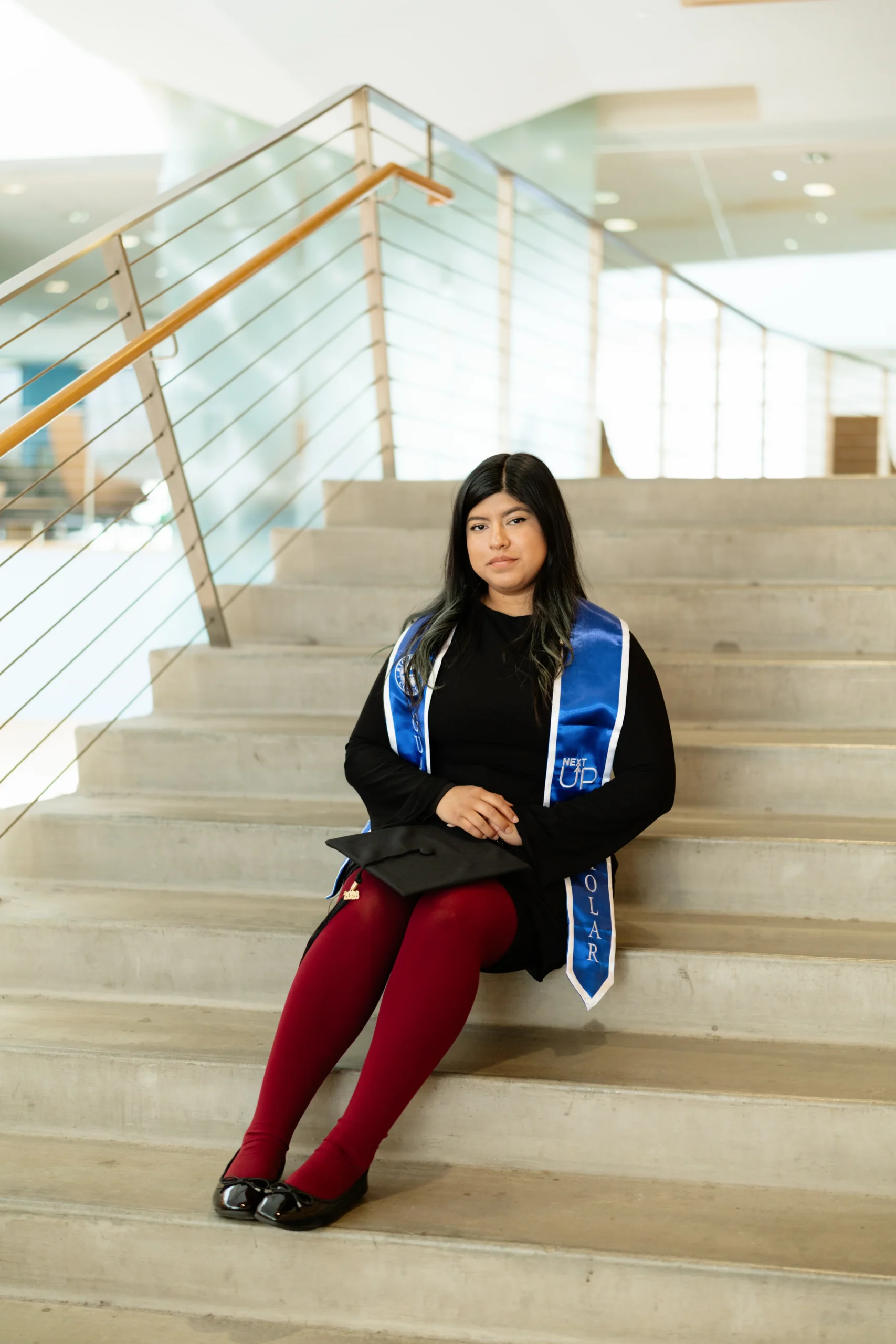 A woman wearing a graduation stole sits on indoor stairs, facing the camera with hands folded in her lap.