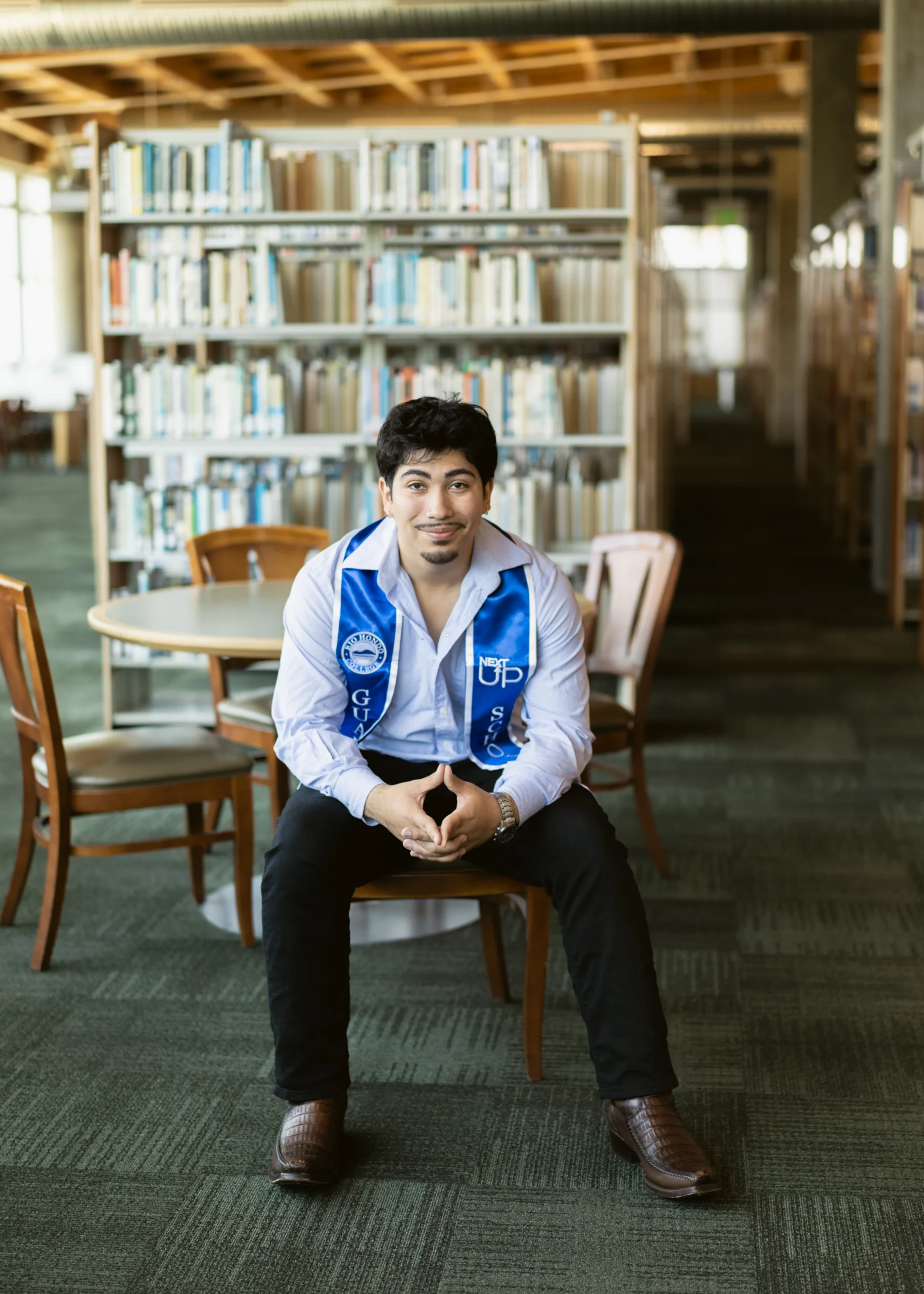 A young man wearing a blue graduation stole sits on a chair in a library, with bookshelves and tables visible in the background.