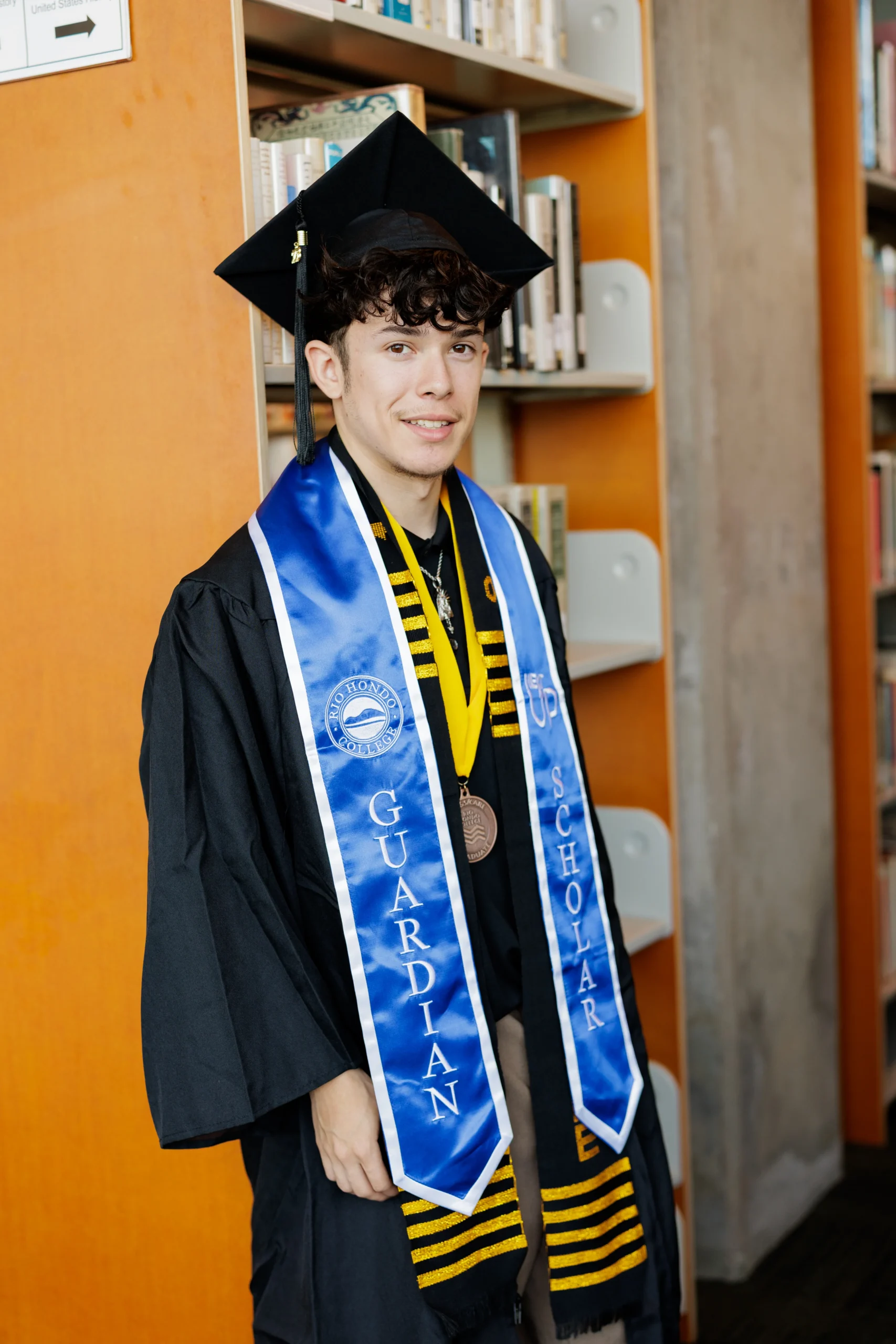 A young person in graduation attire stands in a library, wearing a blue "Guardian Scholar" sash, a medal, and a mortarboard cap.