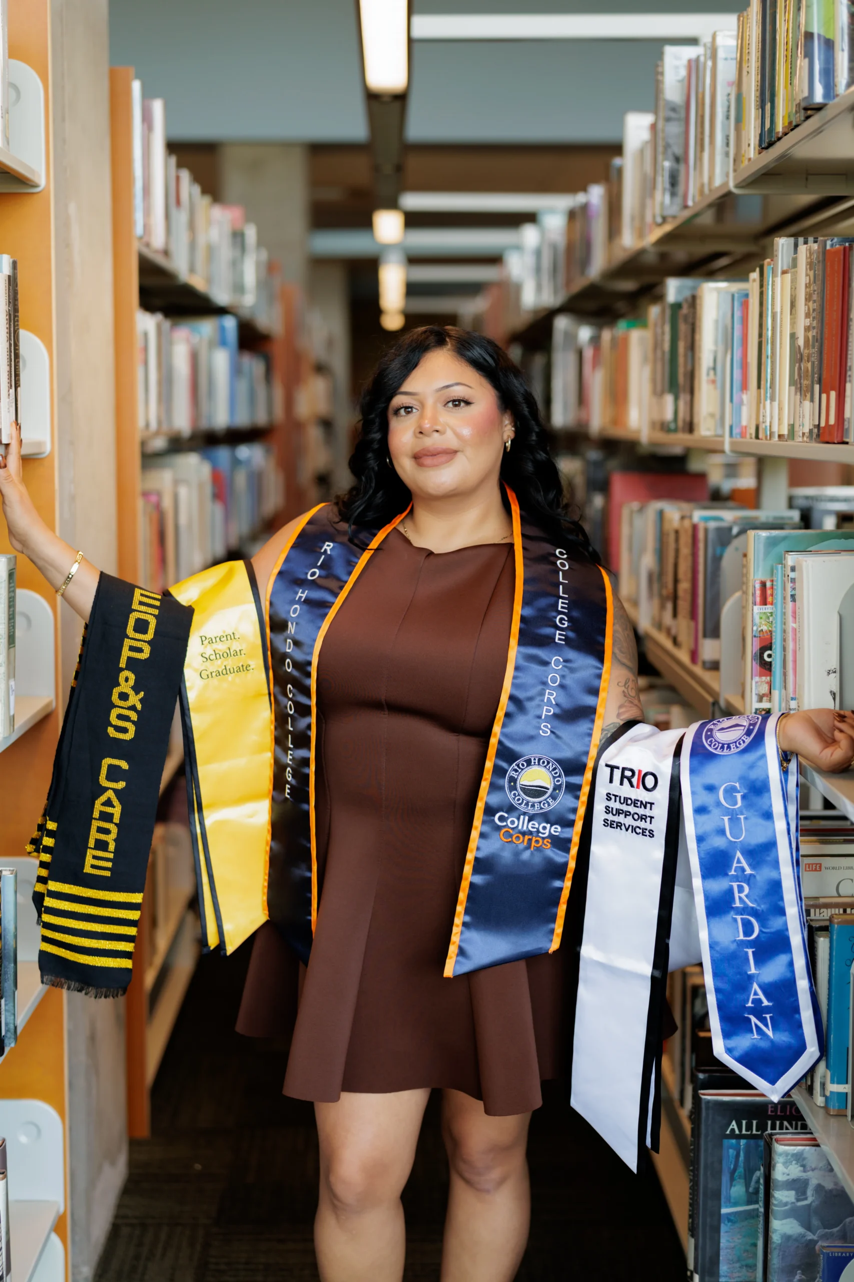 A woman in a brown dress stands between library shelves, wearing multiple graduation stoles and sashes representing academic achievements and programs.