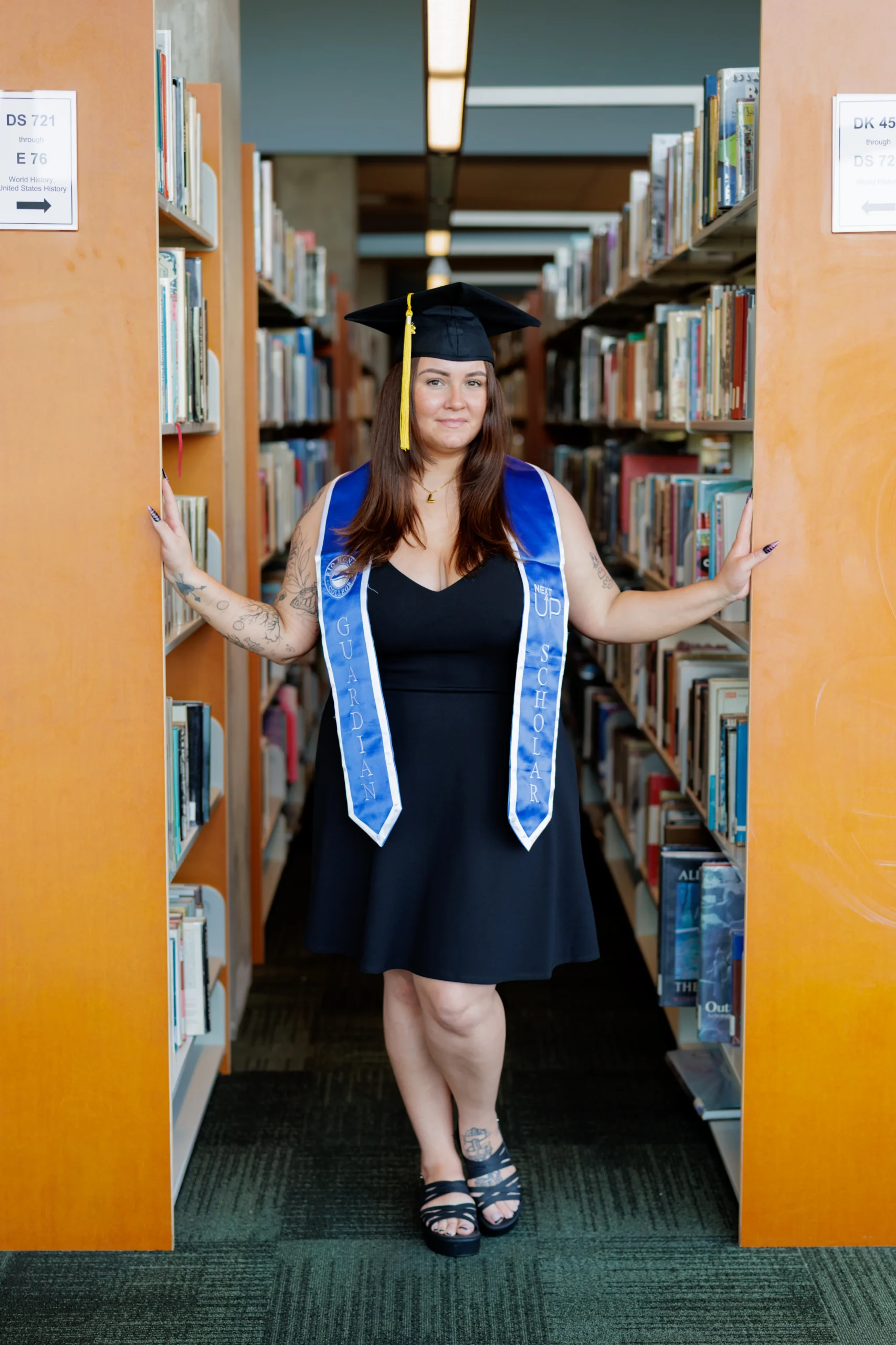 A woman wearing a graduation cap and blue stole stands between bookshelves in a library, dressed in a black dress and sandals.