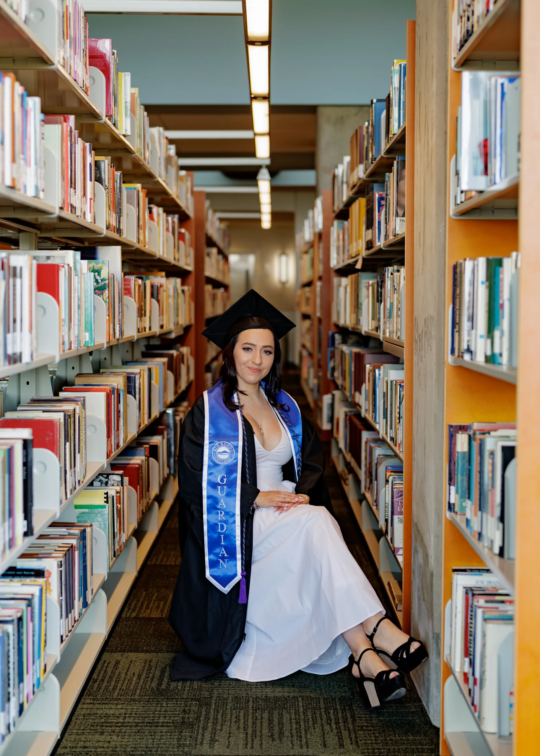 A woman in a graduation cap and gown sits on the floor between bookshelves in a library, wearing a white dress and black heels.