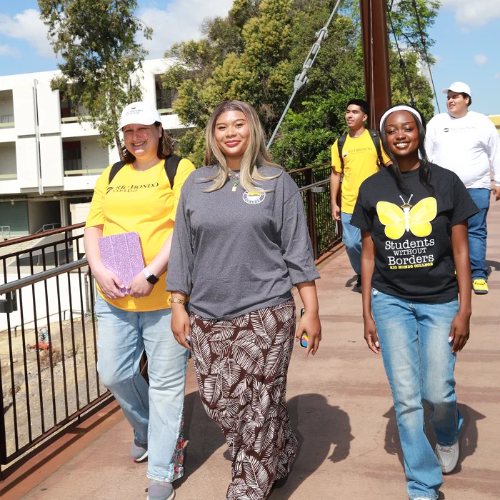 A group of students walks across a bridge on a sunny day, some wearing "Students Without Borders" shirts and carrying backpacks and folders.