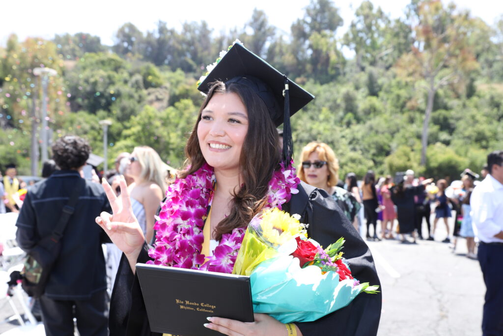 A graduate in a cap and gown holds a diploma and bouquet, smiling and making a peace sign, with people and greenery in the background.