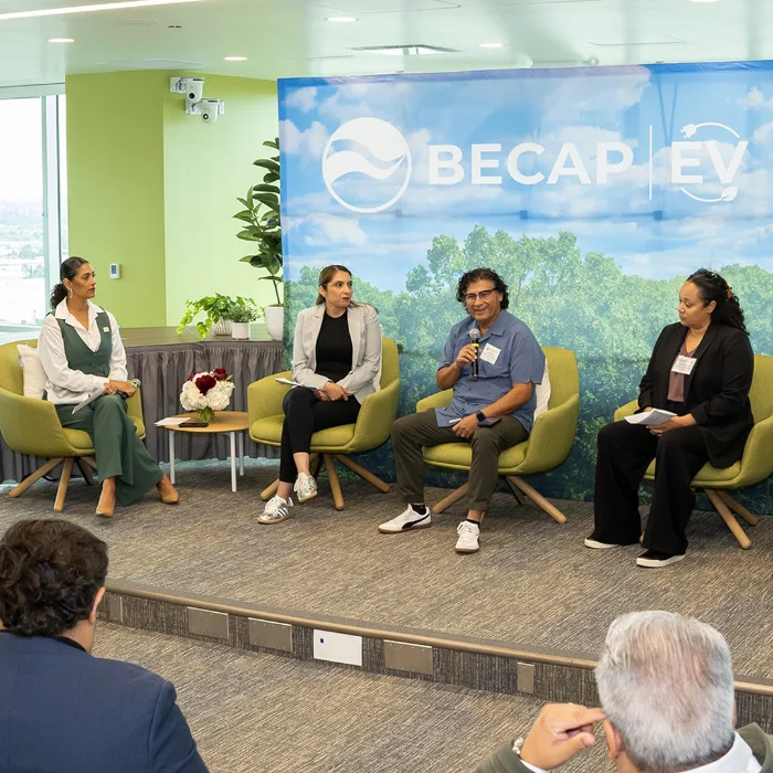 Four people sit on a stage panel discussing at a BECAP EV Summit event, with an audience in the foreground. A banner with a nature motif is displayed behind them.