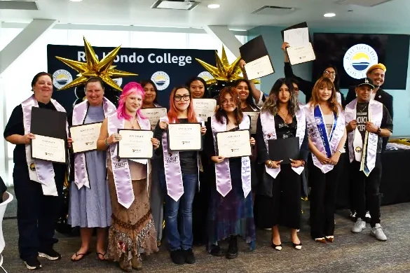 A group of graduates in sashes hold certificates and pose for a photo at a Redondo College ceremony; some wear star-shaped hats.