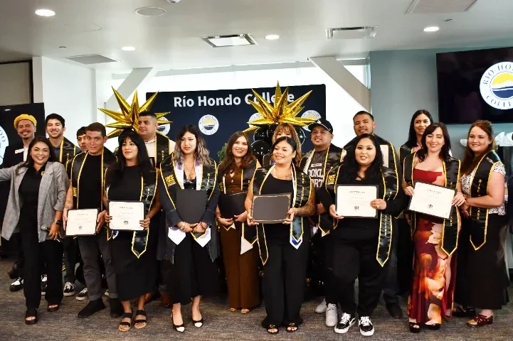 A group of graduates in caps and gowns pose indoors, holding certificates at a Rio Hondo College event. Some wear gold star headpieces.
