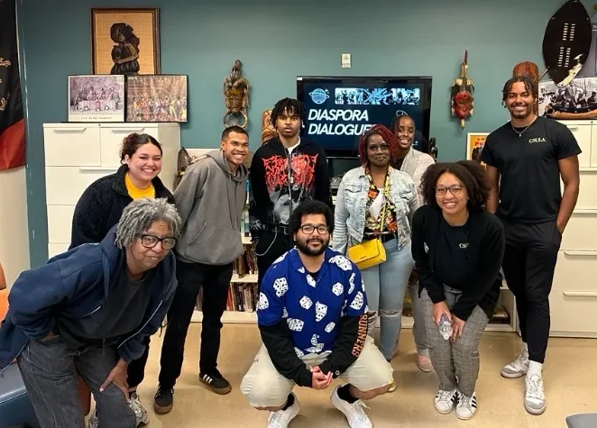 Nine people pose and smile in an office with art and photos on the walls. A monitor behind them displays the words “Diaspora Dialogues.”.