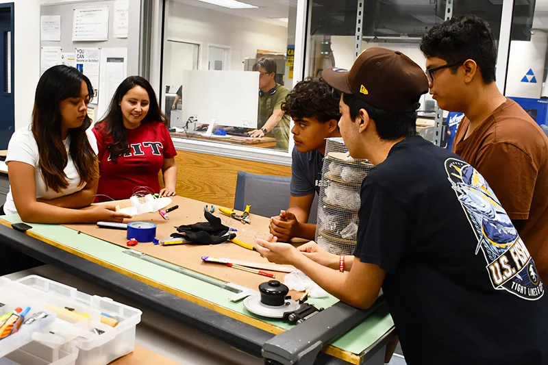 Five students sit and stand around a work table covered with tools and materials, collaborating on a project in a classroom or lab setting.