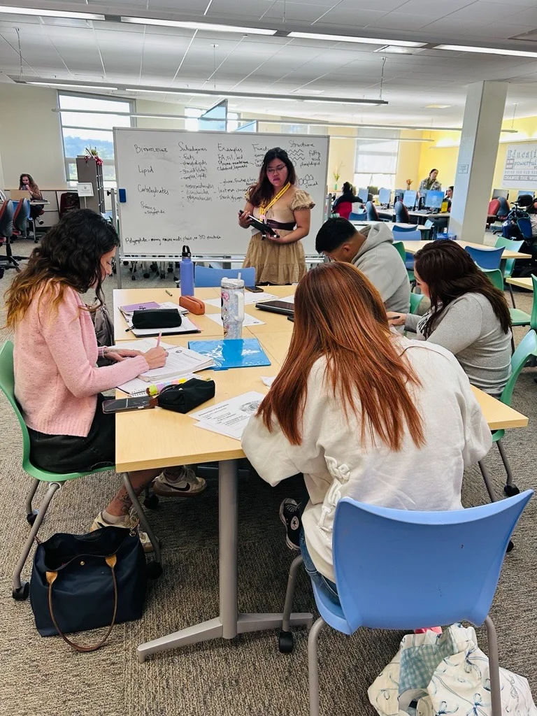 An instructor stands by a whiteboard explaining biochemistry notes to a group of students seated at a table.